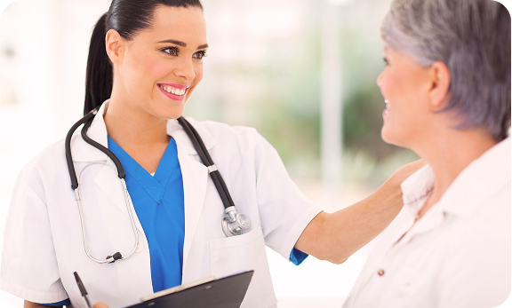 A female doctor in scrubs and a white coat, holding a clipboard, smiles and talks to an older female patient.