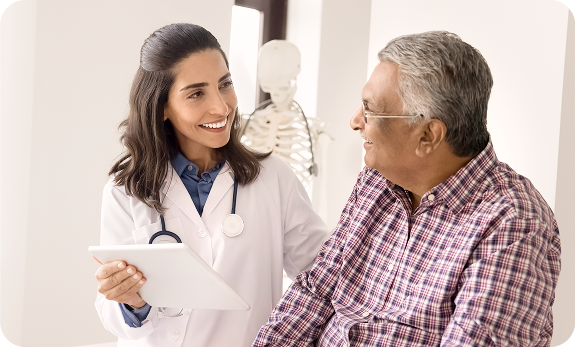 A doctor holding a tablet smiles while talking to an older male patient in a plaid shirt during a medical consultation.
