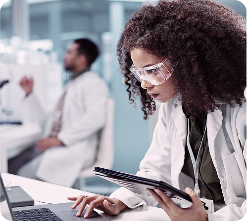 Young woman in lab coat and safety glasses using laptop in laboratory setting.