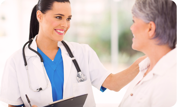 A female doctor in scrubs and a white coat, holding a clipboard, smiles and talks to an older female patient.