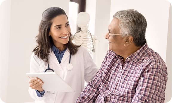 A doctor holding a tablet smiles while talking to an older male patient in a plaid shirt during a medical consultation.
